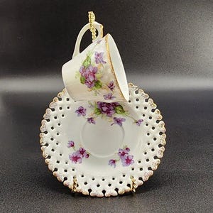 May include: A white teacup and saucer set with a floral violet design and gold trim. The saucer has a delicate, openwork design around the edge. The teacup is suspended above the saucer by a gold-colored stand, creating an elegant display.