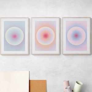 Op de afbeelding: Drie ingelijste abstracte kunstprints met een verloop van roze, blauwe en gele kleuren. Elke print heeft een cirkel met een roze centrum en een witte omtrek.