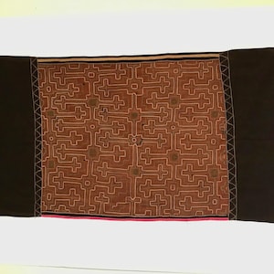 May include: A rectangular textile with a central brown panel featuring a geometric cross pattern in white. The panel is bordered by black fabric on each side. A pink stripe accents the bottom edge.