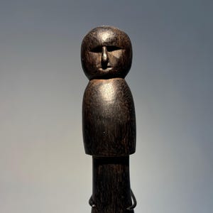 Puede incluir: Una figura de madera tallada de color marrón oscuro, con una cabeza redondeada y una cara sencilla. La figura tiene un cuerpo cilíndrico y una base. La veta de la madera es visible y la figura parece hecha a mano. La figura es un objeto decorativo.