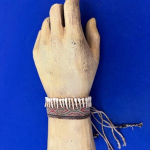 May include: A plaster hand model wearing a bracelet made of small white teeth, a woven band with red, green, and black stripes, and a long tassel of beige string.