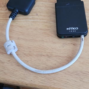 May include: A black NYKO brand adapter with a white cable, connecting two black rectangular connectors. The adapter has the text "NYKO 1234" printed on it. The device is on a wooden surface.
