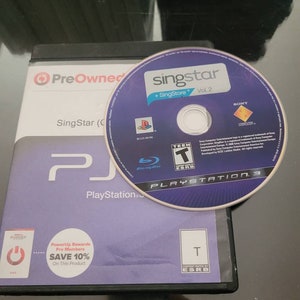 May include: A PlayStation 3 game case and disc for SingStar + SingStore Vol. 2. The disc is purple with white text and the game case is black and purple. The case has the PlayStation logo and the ESRB rating.