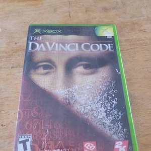 May include: Xbox video game case with a green background and the title "The Da Vinci Code" in white text. The cover features a close-up of a man's face with a blurred background of numbers and symbols.