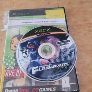 May include: A black Xbox game case with a green and black disc inside. The case has a picture of a cartoon character on the front and the text "Operation Flashpoint: Elite <XBOX>" on the front. The disc has a picture of a soldier in a helmet and the text "Operation Flashpoint Elite" on the front.