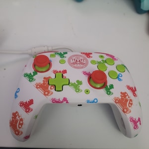 May include: White wireless game controller with a Mario Kart themed design. The controller has green, red, and orange accents. The controller has a logo that says "1992" and "Super Mario Kart".