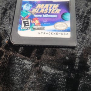 May include: A Nintendo DS game cartridge for "Math Blaster in the Prime Adventure." The game case features a colorful graphic with a cartoon character, a planet, and the game's title. The ESRB rating is "Everyone."