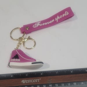 May include: A pink sneaker keychain with the Adidas logo, attached to a pink strap that reads "Forever sports" in white. The keychain has a gold-colored ring and clasp. A ruler is in the foreground.