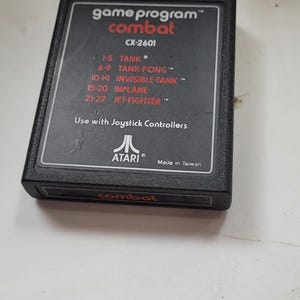 May include: A black rectangular Atari game cartridge for the Combat game program CX-2601. The label lists game modes including Tank, Tank-Pong, Invisible-Tank, Biplane, and Jet-Fighter, with the text Use with Joystick Controllers and Made in Taiwan.