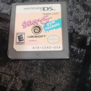 May include: Nintendo DS game cartridge for Ener-G Gym Rockets. The game cartridge has a light pink background with the game title in pink and blue. The game is rated "Everyone" by the ESRB.