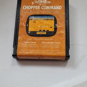 May include: A black Atari video game cartridge for Chopper Command by Activision. The orange label features the game title, a screenshot of the gameplay, and text detailing Cadet and Commander levels, player options, and 1982 copyright info.