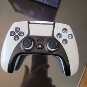 May include: A white and black video game controller with a light blue glow around the buttons. The controller has a home button, a touchpad, and a speaker.