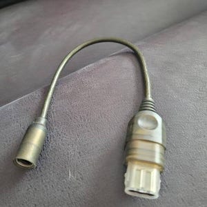 Puede incluir: Un cable negro y gris con un conector en cada extremo. El conector de la izquierda es una toma de audio estándar, y el conector de la derecha es un conector rectangular más grande.