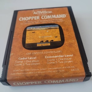 May include: A vintage Atari video game cartridge for the Atari Video Computer System. The cartridge is brown and black with the title "CHOPPER COMMAND" printed in white. The game is by Activision and was designed by Bob Whitehead. The cartridge has a small screen showing a helicopter flying over a desert landscape. The text on the cartridge also includes the game's level options: Cadet Level and Commander Level.