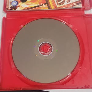 Puede incluir: Una caja de juego roja con un disco plateado y una car&aacute;tula. La car&aacute;tula muestra a una persona con equipo t&aacute;ctico y el t&iacute;tulo del juego "Tom Clancy's Rainbow Six Vegas". El disco tiene un centro rojo.