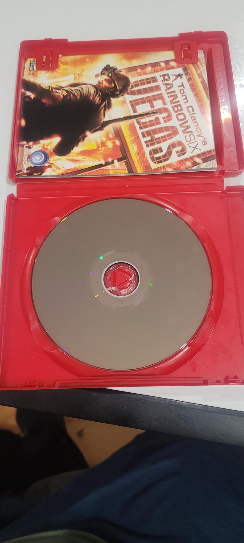 Puede incluir: Una caja de juego roja con un disco plateado y una car&aacute;tula. La car&aacute;tula muestra a una persona con equipo t&aacute;ctico y el t&iacute;tulo del juego "Tom Clancy's Rainbow Six Vegas". El disco tiene un centro rojo.