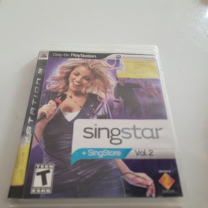 May include: A PlayStation 3 game case for SingStar Vol. 2 featuring a woman singing into a microphone. The game case is white with a purple background and the text "SingStar + SingStore Vol. 2" in blue and white. The game is rated T for Teen.