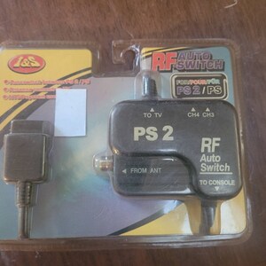 May include: A black RF Auto Switch for PS2/PS consoles. The switch has a black plastic housing with a silver label that reads "PS 2" and "RF Auto Switch". The switch has two inputs, one for the antenna and one for the console, and two outputs, one for the TV and one for the console.
