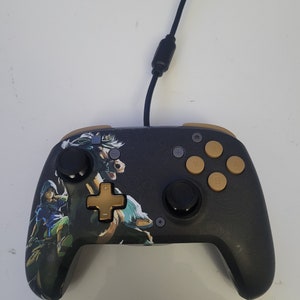 May include: A black and gold video game controller with a Zelda: Breath of the Wild design. The controller has a black and gold d-pad, black and gold buttons, and a black and gold analog stick.
