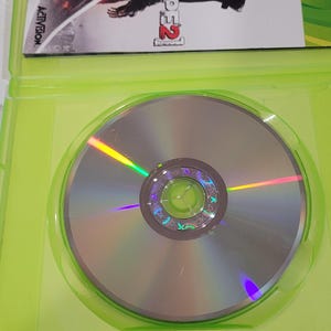 K&ouml;nnte beinhalten: Ein gr&uuml;nes Xbox 360-Spielecase mit einer silbernen Disc im Inneren. Die Vorderseite des Cases zeigt ein rotes und schwarzes Bild eines Charakters aus dem Videospiel Prototype 2. Der Spieltitel "Prototype 2" ist in Wei&szlig; auf der Vorderseite des Cases geschrieben.