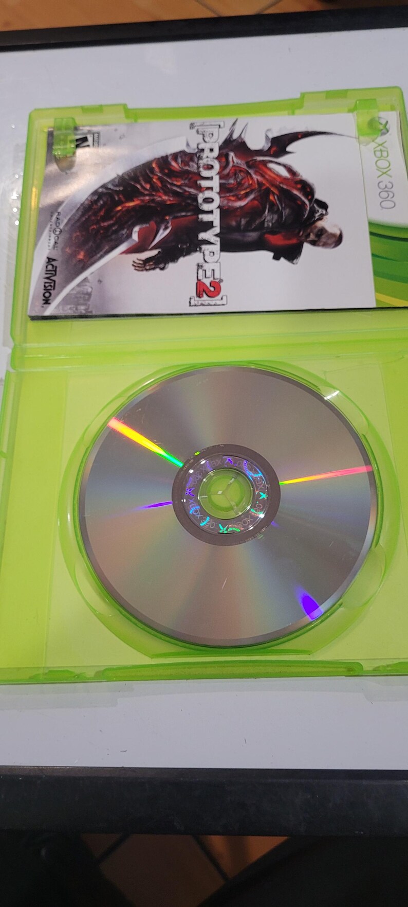 K&ouml;nnte beinhalten: Ein gr&uuml;nes Xbox 360-Spielecase mit einer silbernen Disc im Inneren. Die Vorderseite des Cases zeigt ein rotes und schwarzes Bild eines Charakters aus dem Videospiel Prototype 2. Der Spieltitel "Prototype 2" ist in Wei&szlig; auf der Vorderseite des Cases geschrieben.