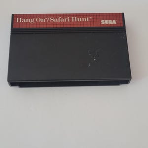 May include: A black Sega Master System game cartridge with a red label that reads "Hang On/Safari Hunt" and "SEGA".