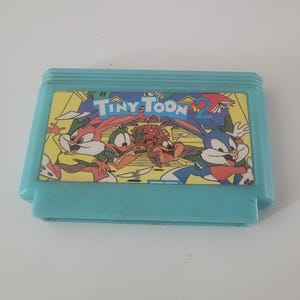 May include: A blue and yellow Nintendo Entertainment System (NES) game cartridge with the title "Tiny Toon 2" printed on the label. The label features cartoon characters from the Tiny Toon Adventures television show.