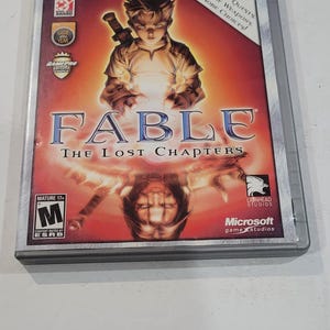 May include: Fable: The Lost Chapters Xbox game case. The cover features a young man with a sword, set against a fiery background. The game case is labeled "Platinum Hits" and includes the ESRB rating "Mature 17+".