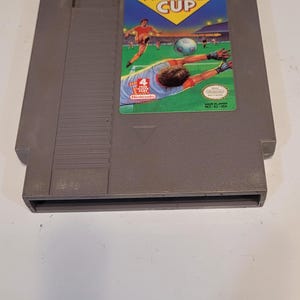 May include: A gray Nintendo Entertainment System (NES) game cartridge for the game "World Cup". The game cartridge features a colorful label with a soccer player kicking a ball and the text "WORLD CUP".