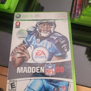 May include: A video game case for Madden NFL 08 for the Xbox 360. The case is green and black with a picture of a football player in a blue and white uniform. The game is rated E for Everyone by the ESRB.