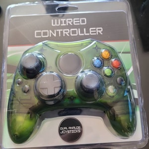May include: A green wired controller for a video game console. The controller has dual analog joysticks and a clear green plastic casing. The packaging says "Wired Controller" and "Dual Analog Joysticks".
