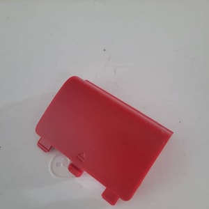 May include: A bright red plastic battery cover with a small triangle symbol on the front.