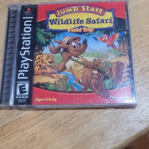 May include: A PlayStation game case for "Jump Start Wildlife Safari: Field Trip." The cover art features a cartoon dog in safari attire, a tiger, and a parrot. The game is rated "Everyone" and for ages 4 and up.