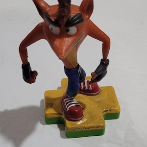 May include: A figurine of Crash Bandicoot, an orange marsupial character, standing on a yellow platform with a green border. He wears blue shorts, red shoes, and black gloves. The figure has a determined expression.