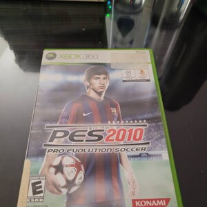 May include: Xbox 360 game case for Pro Evolution Soccer 2010, featuring a soccer player in a red and blue jersey holding a soccer ball. The game case is green and has the text "PES 2010" and "PRO EVOLUTION SOCCER" on it. The game is rated "E" for Everyone by the ESRB.