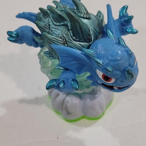 May include: A blue and teal action figure of a dragon-like creature with a spiky back and red eyes. The figure is perched on a cloud-shaped base with green accents. A collectible toy.