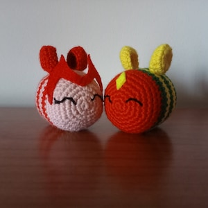 May include: Two crocheted, round ornaments. One is pink with red stripes and a red headpiece. The other is orange with yellow and green stripes and yellow ears. Both have black, smiling faces.