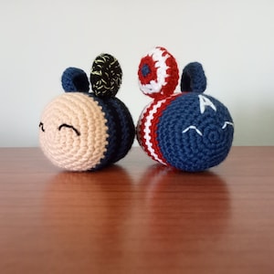 May include: Two crocheted bee plushes, one with a black and yellow striped pattern and the other with a red, white, and blue striped pattern. Both have a smiling face and a small crocheted antenna.