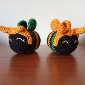 May include: Two crocheted bumblebees with black bodies, green and orange wings, and white smiling faces.