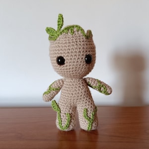 Pode incluir: Boneco Groot de crochê, em fio bege com detalhes verdes. O boneco tem olhos grandes e pretos e folhas verdes na cabeça e nos membros. A figura feita à mão está sobre uma superfície de madeira.