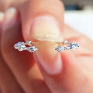 May include: A silver-toned open ring featuring a series of small, clear, round-cut stones set along the band. The metal band has a polished finish and the text RHDSTYLEJ is visible in the centre gap between the two ends of the open ring design.