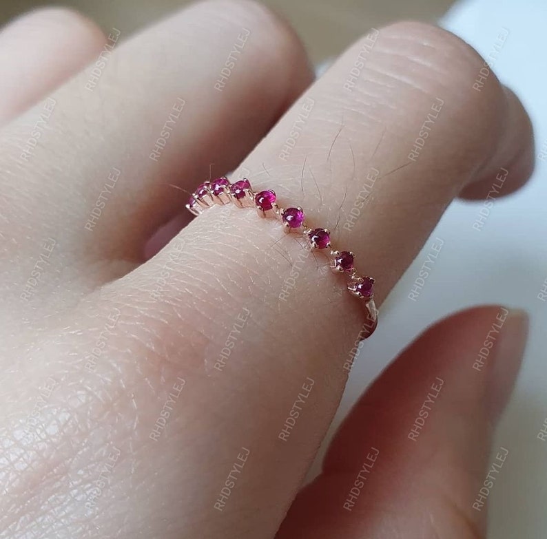 May include: A delicate gold ring with a row of small, bright red gemstones. The ring is worn on a finger.