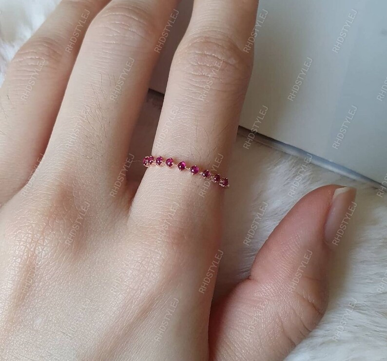 May include: A delicate gold ring with a row of small, deep red gemstones. The ring is worn on a finger.