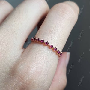 May include: A delicate gold band ring with a row of small, red gemstones. The ring is on a finger.