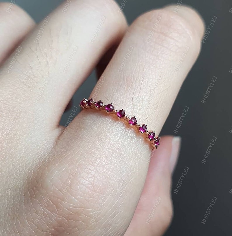 May include: A delicate gold band ring with a row of small, red gemstones. The ring is on a finger.