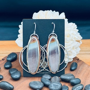 May include: A pair of handcrafted earrings with oval-shaped, multi-colored stone insets. The stones are framed in silver and suspended from silver hoops. The earrings are displayed on a black card with the text "Kiki Handmade Jewelry".
