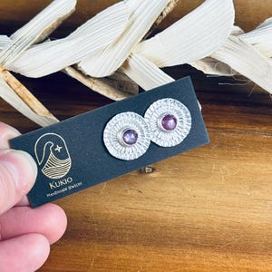 May include: A pair of silver earrings with a textured circular design and a small purple gemstone in the center. The earrings are displayed on a black card with the text "KUKIO HANDMADE JEWELRY".