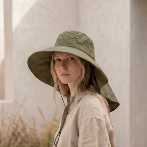 May include: A woman wearing a wide-brimmed olive green sun hat with a chin strap. She is wearing a beige linen shirt and white pants. The background features a neutral-toned architectural setting with tall, rectangular structures and tall grass.