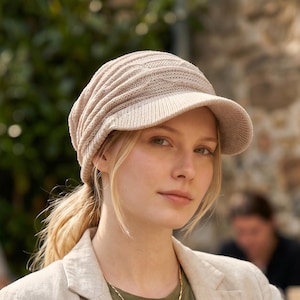 Può includere: Una donna indossa un berretto di maglia beige con visiera, un blazer color beige chiaro e una maglietta verde. È visibile una collana a catena dorata. Lo sfondo include un muro di pietra e vegetazione.
