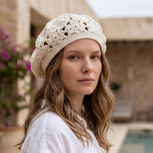 May include: A cream-colored crochet beret featuring a detailed floral pattern throughout the crown and a solid, tight-knit band around the base. The headwear is crafted from yarn with a soft texture and a consistent off-white color tone.